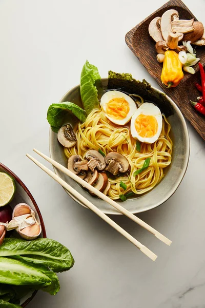 Vista superior de ramen asiático tradicional em tigela perto de pauzinhos, legumes na superfície cinza — Fotografia de Stock