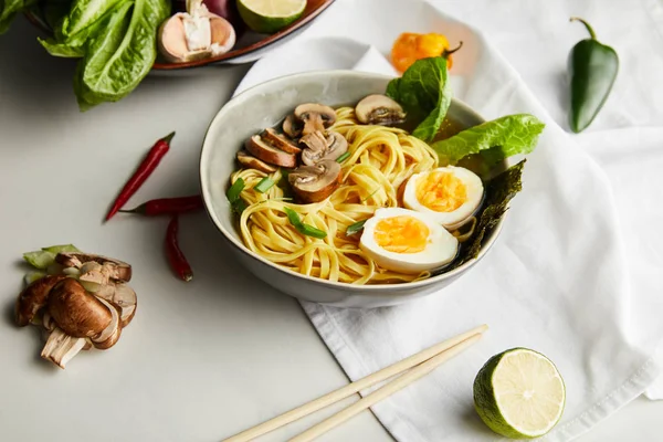 Традиционный азиатский рамен в миске возле палочек для еды, салфеток и овощей на серой поверхности — стоковое фото
