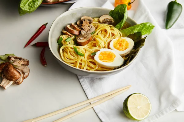 Tradicional asiático ramen em tigela perto de pauzinhos, guardanapo e legumes na superfície cinza — Fotografia de Stock