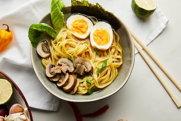 Top view of traditional asian ramen in bowl near chopsticks, napkin and vegetables on grey surface — Stock Photo