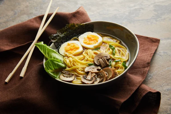 Traditional spicy ramen in bowl with chopsticks on brown napkin on stone surface — Stock Photo