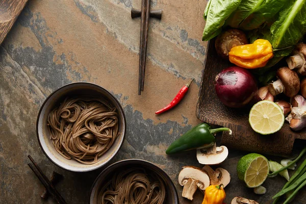 Vista superior de macarrão picante tradicional em tigelas perto de pauzinhos e legumes frescos na superfície de pedra — Fotografia de Stock