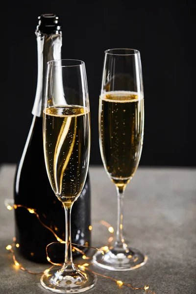 Gläser und Flasche mit Sekt und gelber Weihnachtsbeleuchtung, auf schwarz — Stockfoto