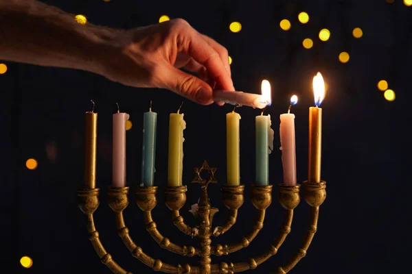 Vista cortada do homem acendendo velas em menorah no fundo preto com luzes bokeh em Hanukkah — Fotografia de Stock