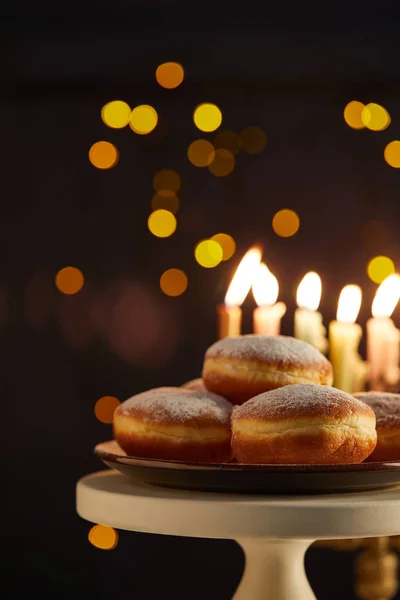 Selective focus of delicious doughnuts on stand near glowing candles on black background with bokeh lights on Hanukkah — Stock Photo