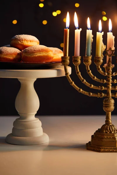 Deliciosos donuts em stand perto de velas brilhantes em menorah no fundo preto com luzes bokeh em Hanukkah — Fotografia de Stock