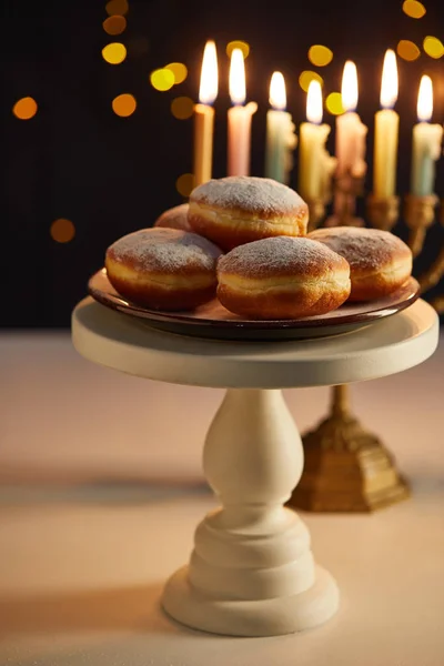Foco seletivo de deliciosos donuts em stand perto de velas brilhantes em menorah no fundo preto com luzes bokeh em Hanukkah — Fotografia de Stock