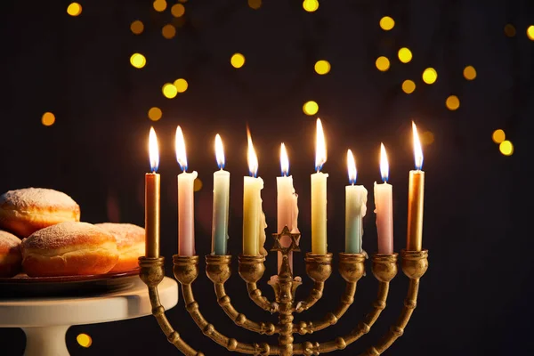 Delicious doughnuts on stand near glowing candles in menorah on black background with bokeh lights on Hanukkah — Stock Photo