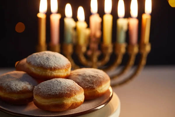 Selective focus of delicious doughnuts on stand near glowing candles in menorah on black background with bokeh lights on Hanukkah — Stock Photo