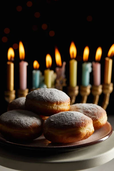 Selective focus of delicious doughnuts on stand near glowing candles in menorah on black background with bokeh lights on Hanukkah — Stock Photo