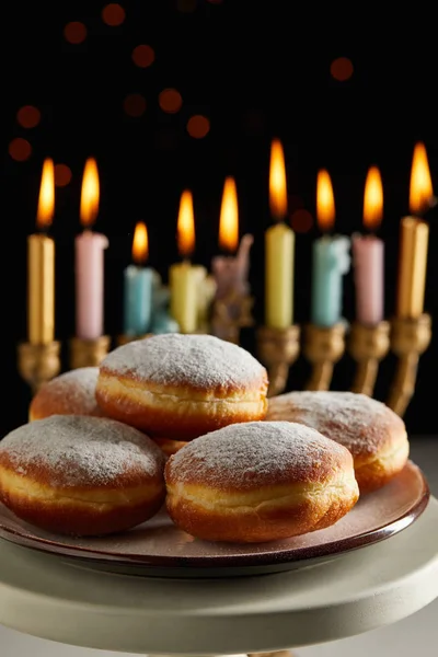 Selective focus of delicious doughnuts on stand near glowing candles in menorah on black background with bokeh lights on Hanukkah — Stock Photo