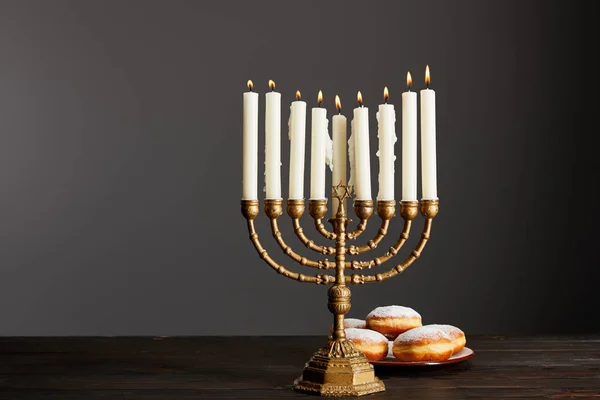 Delicious doughnuts near burning candles in menorah on wooden table on Hanukkah on grey — Stock Photo