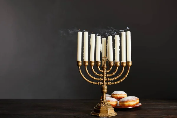 Delicious doughnuts near candles in menorah on wooden table on Hanukkah on grey — Stock Photo