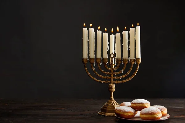 Delicious doughnuts near burning candles in menorah on Hanukkah isolated on black — Stock Photo