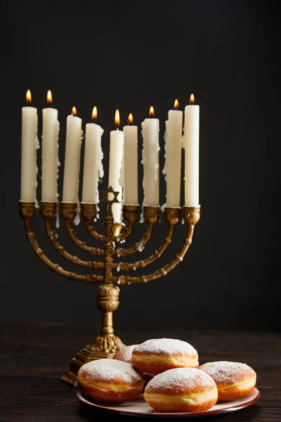 Delicious doughnuts near burning candles in menorah on Hanukkah isolated on black — Stock Photo