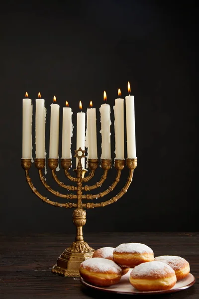 Delicious doughnuts near burning candles in menorah on Hanukkah isolated on black — Stock Photo
