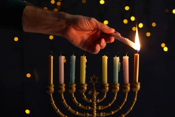 Partial view of man lighting up candles in menorah on black background with bokeh lights on Hanukkah — Stock Photo