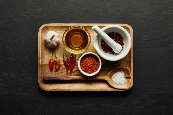Vista dall'alto di spezie, aglio e olio d'oliva su un piatto di legno su sfondo nero — Foto stock
