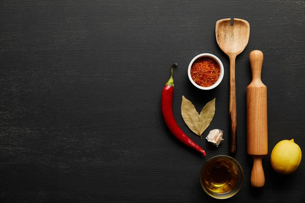Top view of wooden rolling pin and spoon with spices and olive oil on black wooden background — Stock Photo