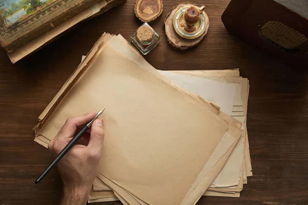 Vista cortada do homem segurando caneta-tinteiro acima do papel vintage perto de pintura, vela e rádio velho na mesa de madeira — Fotografia de Stock