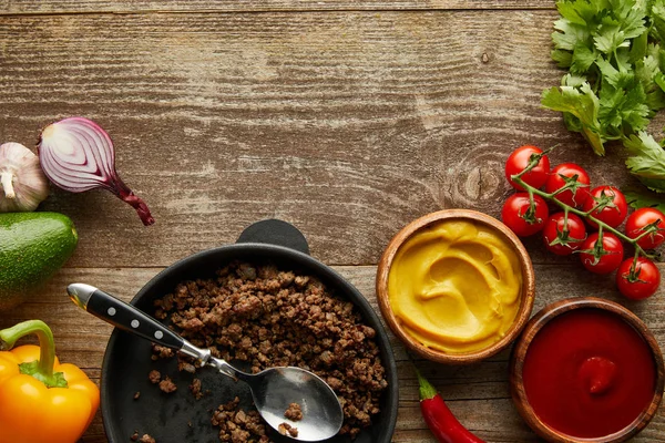Вид сверху фарша в сковороде с соусами и сырыми овощами на деревянном фоне — стоковое фото