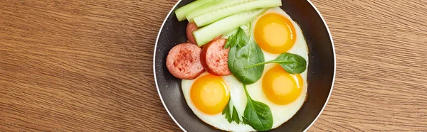 Top view of fried eggs with spinach leaves, cucumber and sausage in frying pan on wooden table, panoramic shot — Stock Photo