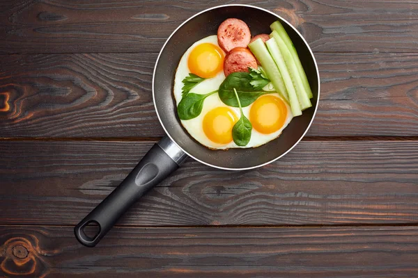 Top view of fried eggs with spinach leaves, cucumber and sausage in frying pan on wooden table — Stock Photo