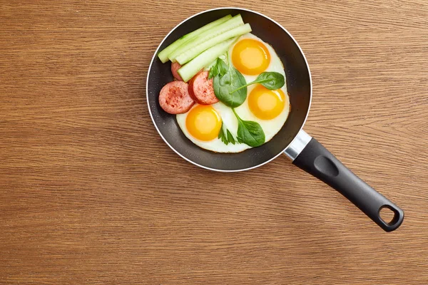 Top view of fried eggs with spinach leaves, cucumber and sausage in frying pan on wooden table — Stock Photo