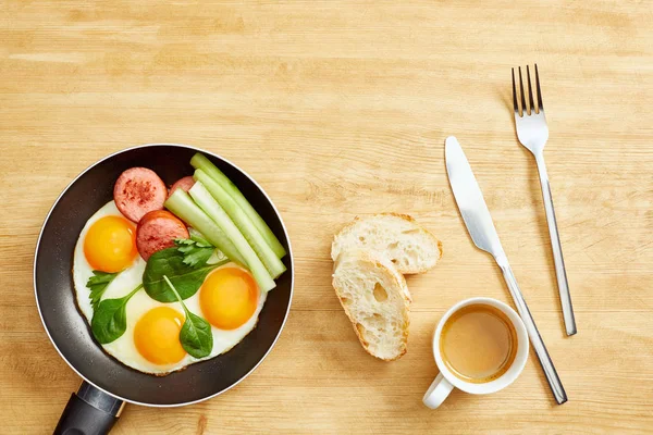 Top view of fried eggs with spinach leaves, cucumber and sausage in frying pan near coffee, bread and coffee on wooden table — Stock Photo