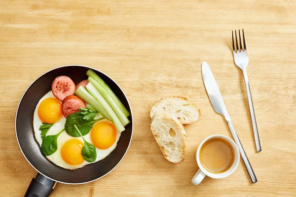 Vista superior de ovos fritos com folhas de espinafre, pepino e salsicha na frigideira perto de café, pão e café na mesa de madeira — Fotografia de Stock