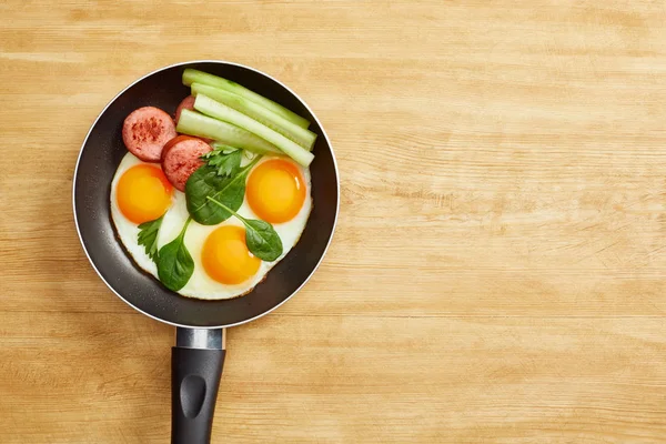 Top view of fried eggs with spinach leaves, cucumber and sausage in frying pan on wooden table — Stock Photo