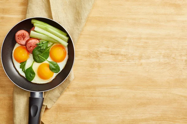 Top view of fried eggs with spinach leaves, cucumber and sausage in frying pan on napkin on wooden table — Stock Photo