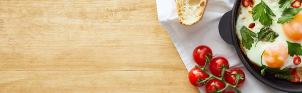 Top view of fried eggs near bread and tomatoes on napkin on wooden table, panoramic shot — Stock Photo