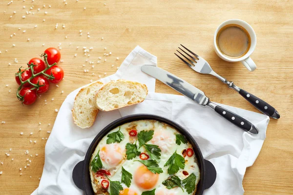 Vista superior de ovos fritos na panela perto de pão, talheres, café e tomates em guardanapo na mesa de madeira — Fotografia de Stock
