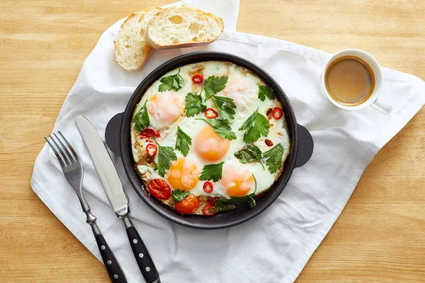 Draufsicht auf Spiegeleier in Pfanne neben Brot, Besteck, Kaffee auf Serviette auf Holztisch — Stockfoto