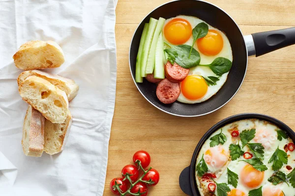 Top view of fried eggs with vegetables in frying pans near napkin with bread on wooden table — Stock Photo