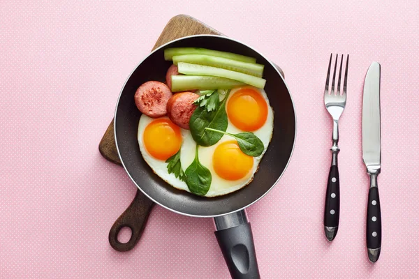 Top view of fried eggs with spinach leaves, cucumber and sausage in frying pan near cutlery on pink dotted background — Stock Photo