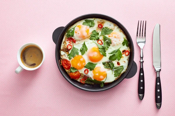 Top view of fried eggs with parsley, tomatoes and chili pepper in frying pan near cutlery and coffee on pink background — Stock Photo