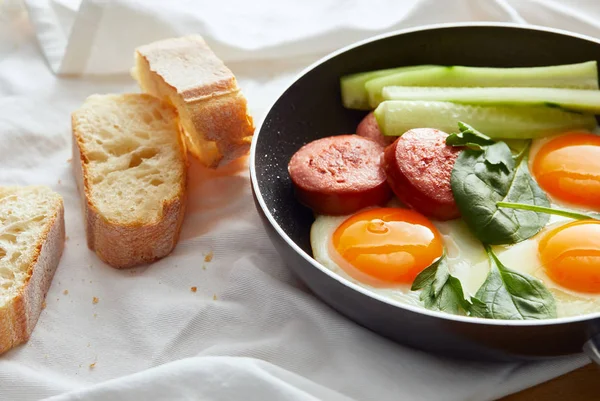 Fried eggs in frying pan with spinach, cucumber and sausage near bread on napkin — Stock Photo