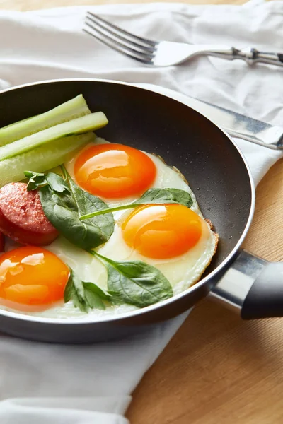 Foco selectivo de huevos fritos en sartén con espinacas, pepino y salchichas en mesa de madera con cubiertos en servilleta - foto de stock