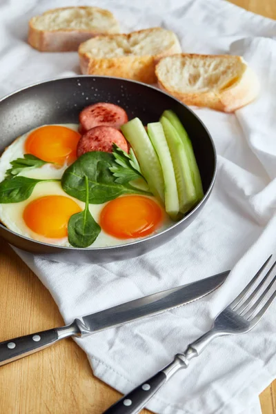 Selective focus of fried eggs in frying pan with spinach, cucumber and sausage at wooden table with cutlery and bread on napkin — Stock Photo