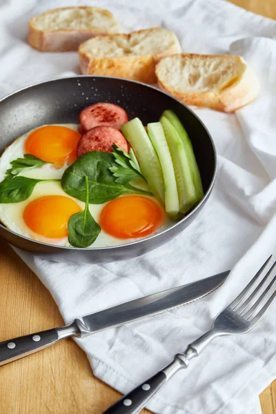 Selektiver Schwerpunkt Spiegeleier in Pfanne mit Spinat, Gurke und Wurst am Holztisch mit Besteck und Brot auf Serviette — Stockfoto