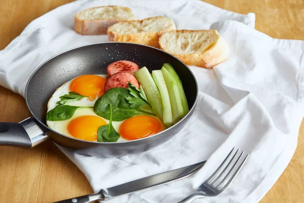 Foyer sélectif des œufs frits dans une poêle aux épinards, concombre et saucisse à table en bois avec couverts et pain sur serviette — Photo de stock