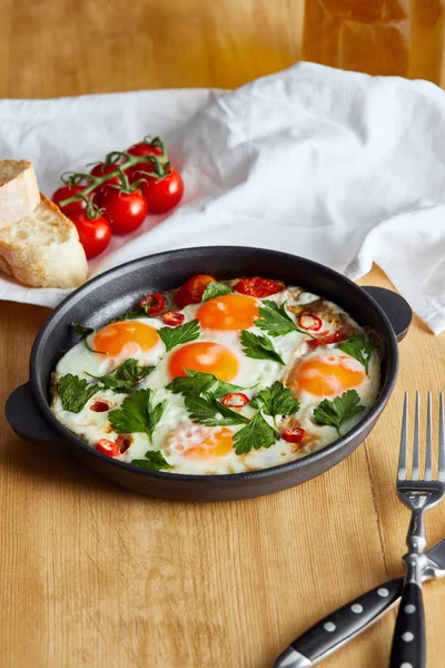 Fried eggs with parsley and chili pepper in pan near napkin, bread, tomatoes and napkin on wooden table — Stock Photo