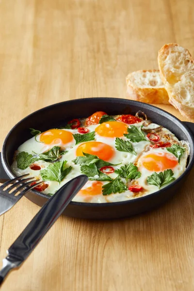 Fried eggs with parsley and chili pepper on wooden table with cutlery and bread — Stock Photo