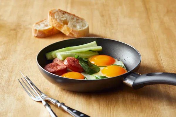 Селективный фокус яиц на сковороде со шпинатом, огурцом и колбасой за деревянным столом со столовыми приборами и хлебом — стоковое фото