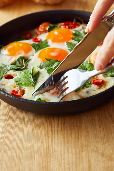 Cropped view of eating fried eggs with parsley and chili pepper on wooden table with fork and knife — Stock Photo