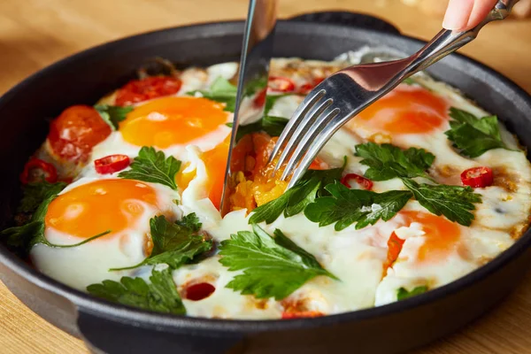 Cropped view of eating fried eggs with parsley and chili pepper on wooden table with fork and knife — Stock Photo