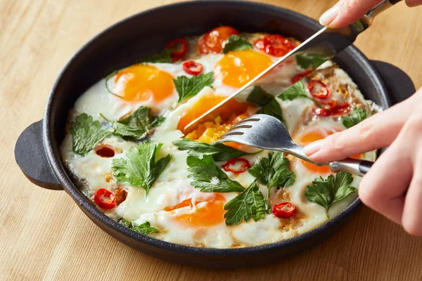 Vista recortada de comer huevos fritos con perejil y chile sobre mesa de madera con tenedor y cuchillo - foto de stock