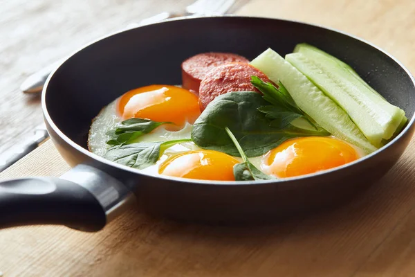 Spiegeleier in der Pfanne mit Spinat, Gurke und Wurst am Holztisch — Stockfoto